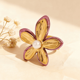 The Orchid Ring, a sculptural gold flower with fuchsia-pink enamel edges and a central white pearl, displayed on a white surface with a rose quartz crystal in the background. The ring is an open, adjustable cuff design.