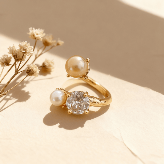 A gold version of The Celestial Ring displayed side-by-side on a neutral, textured brown surface. Each adjustable open ring features one large CZ crystal and two pearls in an asymmetrical design.