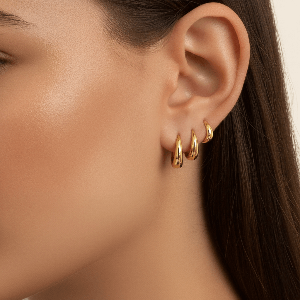A close-up of a model's ear showing a curated ear stack. She is wearing the large gold Capsule Hoop in her first lobe piercing, the medium hoop in her second, and the small hoop in her upper cartilage (helix) piercing.