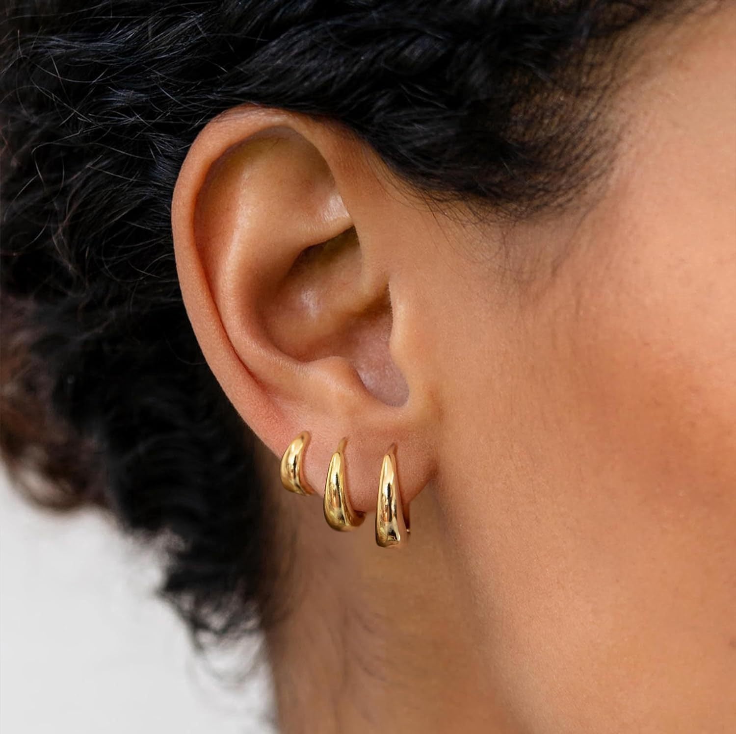 A close-up of a model's ear showing a curated ear stack. She is wearing the large gold Capsule Hoop in her first lobe piercing, the medium hoop in her second, and the small hoop in her upper cartilage (helix) piercing.