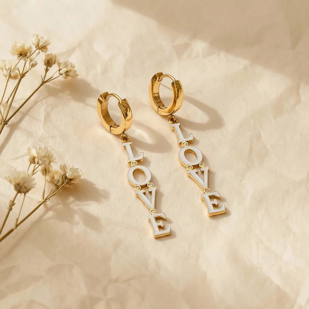 A detailed studio shot of The Amor Earrings by Luminova, featuring the white enamel 'LOVE' letter charms dangling from polished gold huggie hoops, styled with dried flowers.