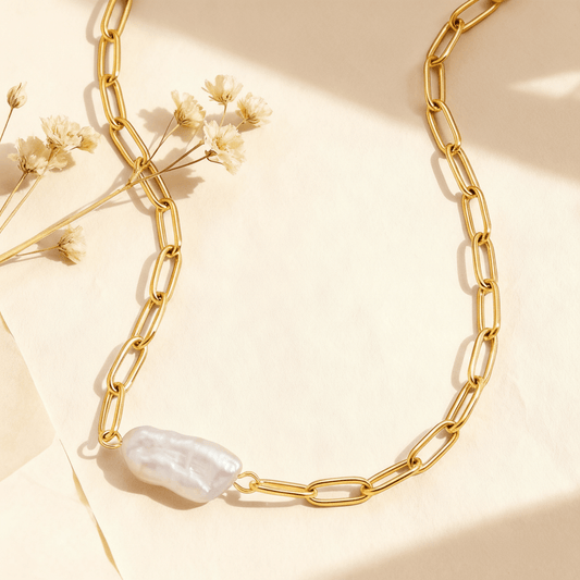 A detailed close-up of The Aphrodite Necklace by Luminova, showcasing the unique, elongated baroque pearl on a waterproof 18k gold-plated paperclip chain, styled on a soft, sunlit background.