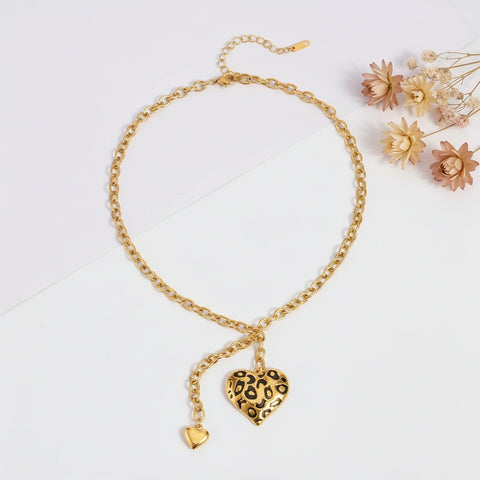 A detailed studio shot of The Artemis Lariat by Luminova, a gold Y-necklace with a bold leopard print heart pendant and a smaller gold heart drop, styled with dried flowers.