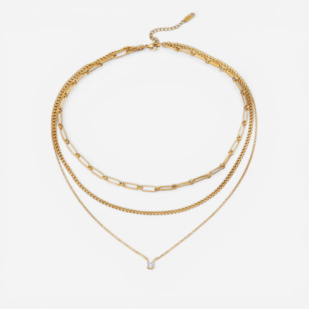 A detailed studio shot of The Charites Necklace by Luminova, showcasing its pre-layered, triple-strand design featuring three unique gold chains and a single sparkling crystal.