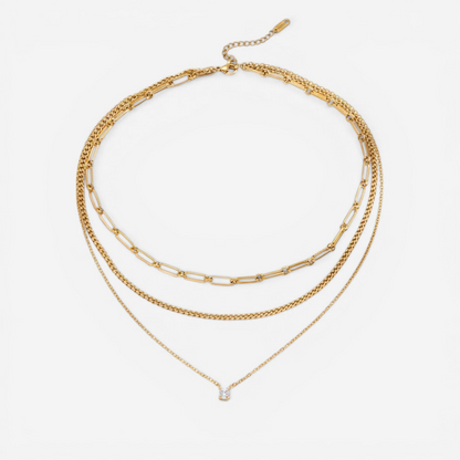 A detailed studio shot of The Charites Necklace by Luminova, showcasing its pre-layered, triple-strand design featuring three unique gold chains and a single sparkling crystal.