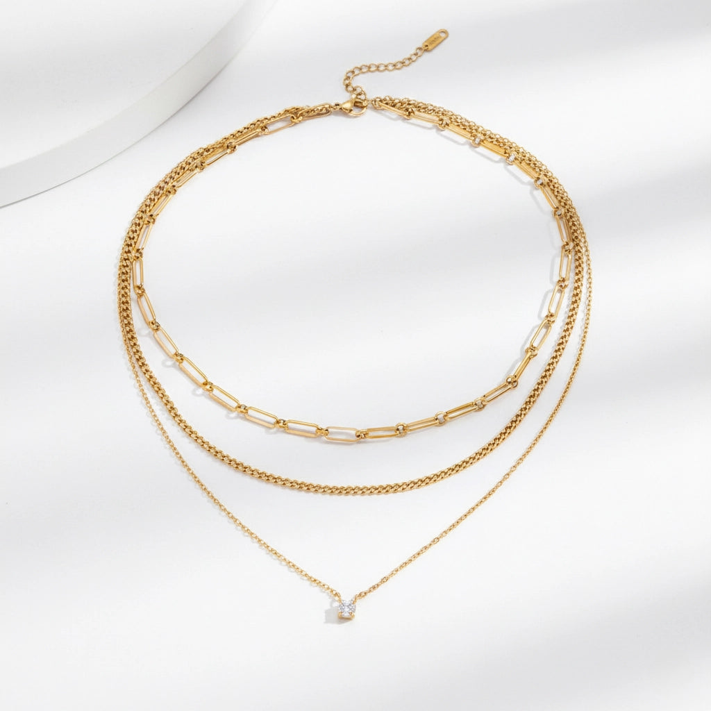 A detailed studio shot of The Charites Necklace by Luminova, showcasing its pre-layered, triple-strand design featuring three unique gold chains and a single sparkling crystal.