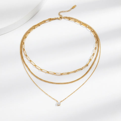 A detailed studio shot of The Charites Necklace by Luminova, showcasing its pre-layered, triple-strand design featuring three unique gold chains and a single sparkling crystal.