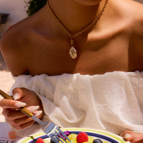 A model wearing The Fleur Pendant with a white off-the-shoulder top in a sunny, beachside setting, embodying effortless summer elegance.