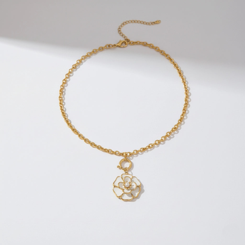 A detailed studio shot of The Fleur Pendant by Luminova, showcasing the white enamel camellia flower on a gold rolo chain, styled in warm sunlight.