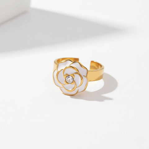A detailed studio shot of The Fleur Ring by Luminova, showcasing the white enamel camellia flower with a sparkling crystal center on an adjustable gold band.
