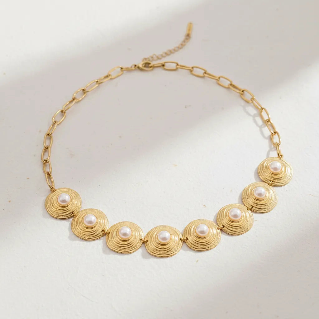 A detailed studio shot of The Hera Necklace by Luminova, showcasing the vintage-style pearl and textured gold medallion collar on a soft, warm background.