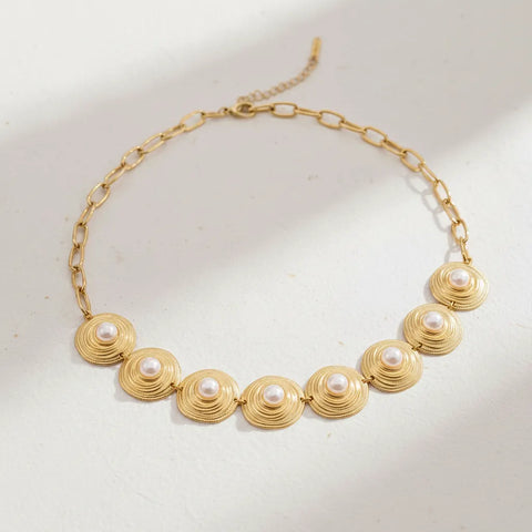 A detailed studio shot of The Hera Necklace by Luminova, showcasing the vintage-style pearl and textured gold medallion collar on a soft, warm background.