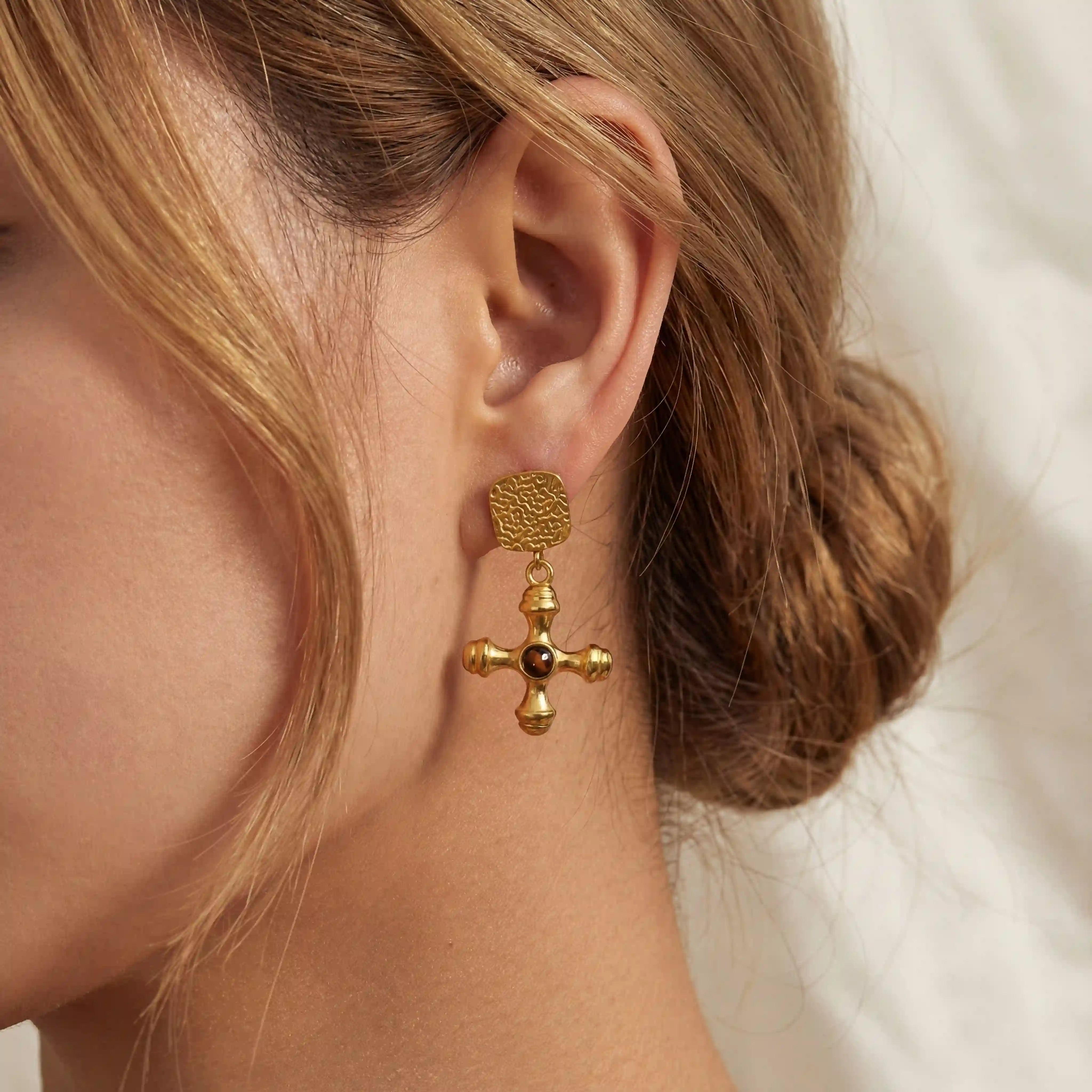 Close up of woman wearing Jupiter gold cross dangle earrings with vintage textured finish. Main image