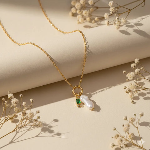 A detailed product shot of The Astoria Necklace for May, highlighting the rich emerald green birthstone charm and lustrous baroque pearl.