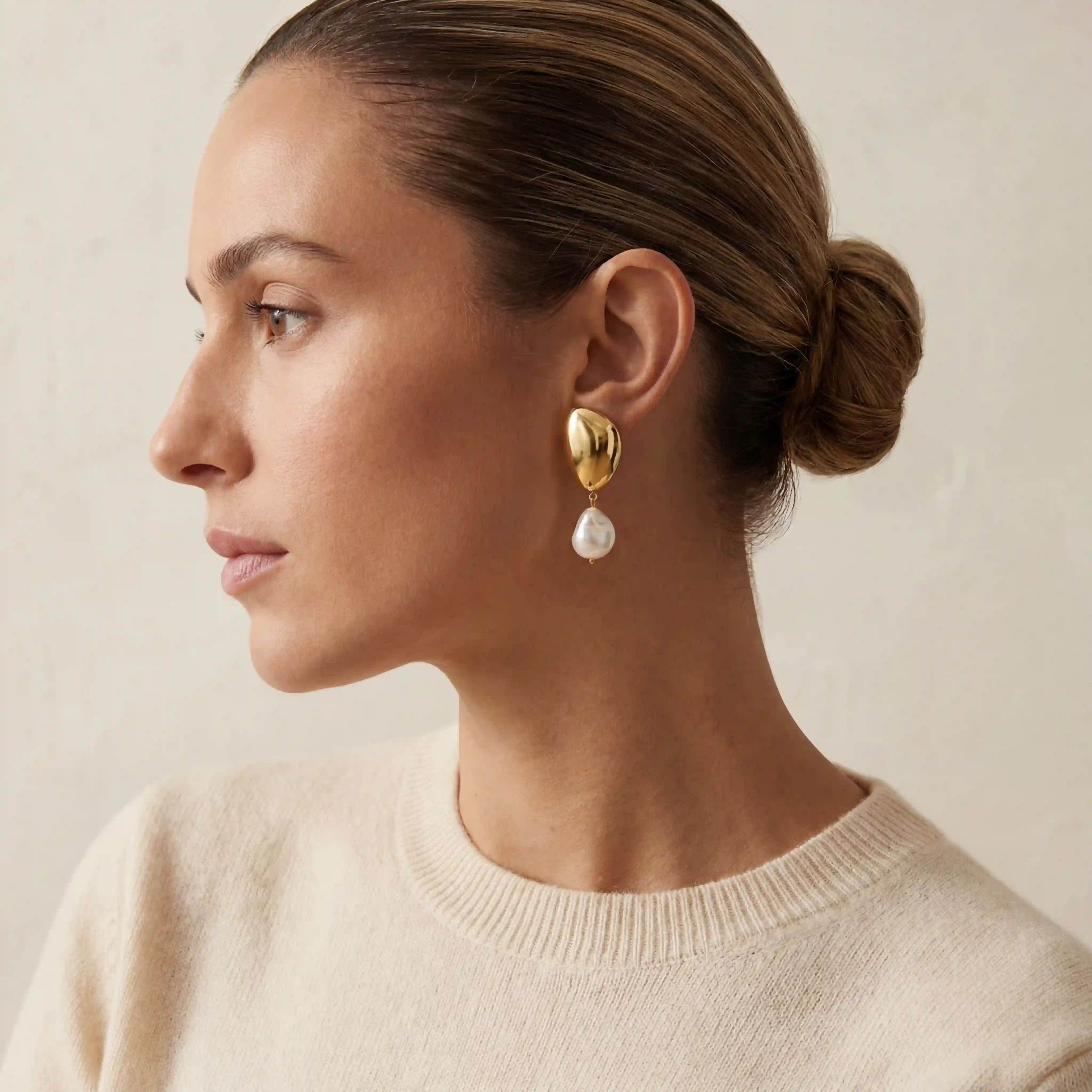 Elegant woman wearing gold statement pearl earrings styled with cream sweater.
