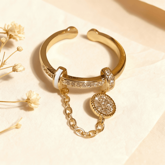 The Comet Ring, an adjustable gold open cuff ring, resting in a white jewelry box. The ring features a dangle chain with a sparkling pavé disc charm, a crystal-lined band, and white enamel accents.