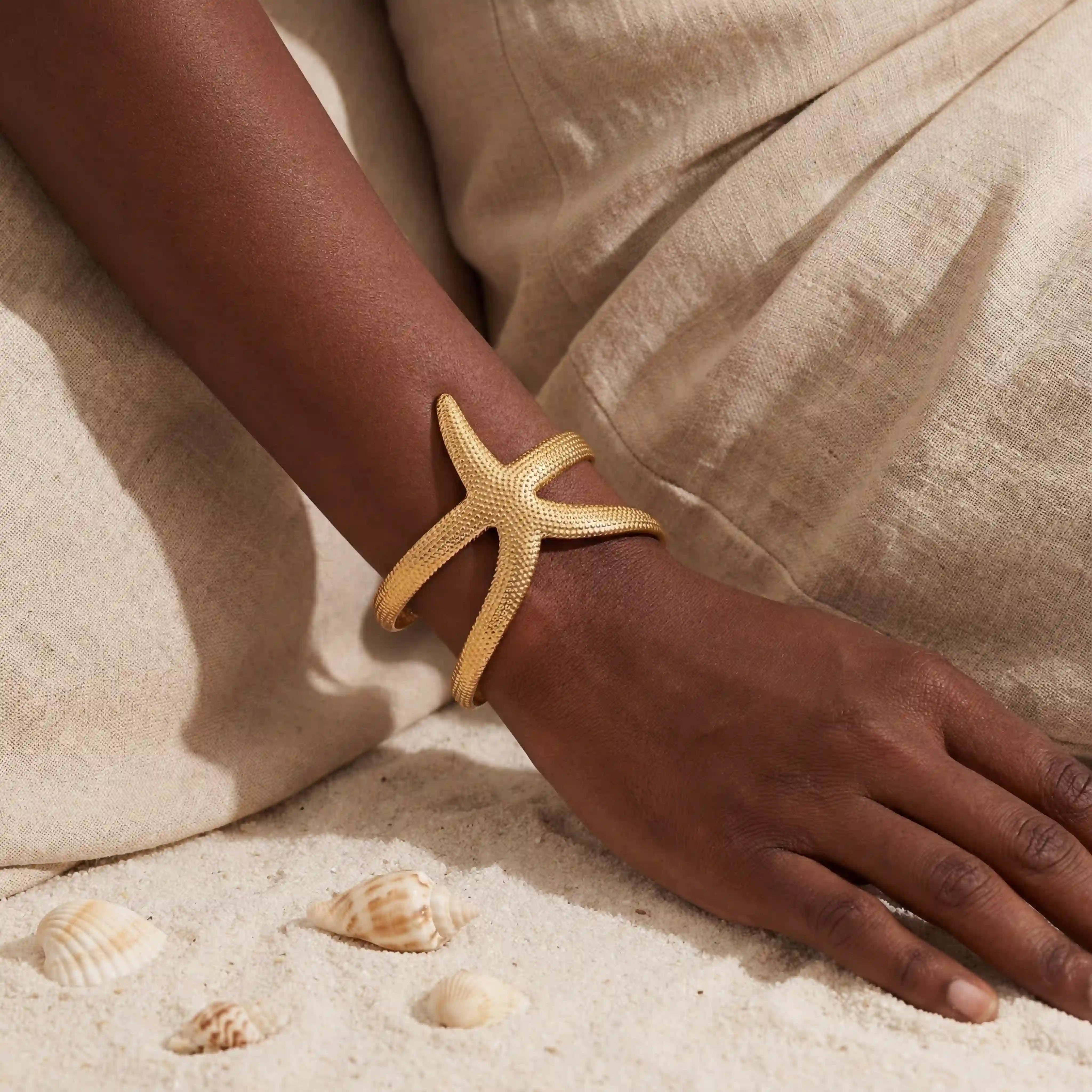 Dark skin model wearing textured gold starfish open cuff bracelet on beach sand summer vibe. Main image