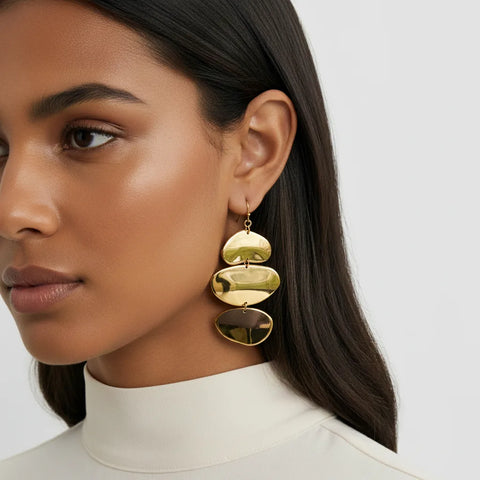 A close-up of a model wearing The Thalia Earrings with a white turtleneck, highlighting their bold scale and how the sculptural gold tiers drop elegantly.