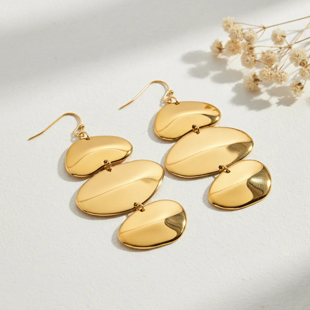 A detailed studio shot of The Thalia Earrings by Luminova, showcasing the cascading design of three organic, polished gold discs, styled with delicate dried flowers.