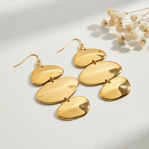 A detailed studio shot of The Thalia Earrings by Luminova, showcasing the cascading design of three organic, polished gold discs, styled with delicate dried flowers.