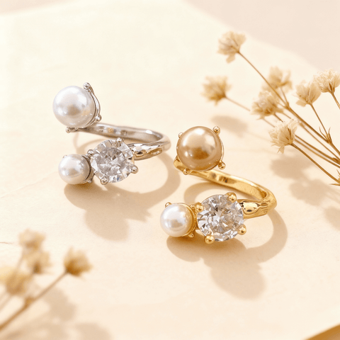 A gold and a silver version of The Celestial Ring displayed side-by-side on a neutral, textured brown surface. Each adjustable open ring features one large CZ crystal and two pearls in an asymmetrical design.