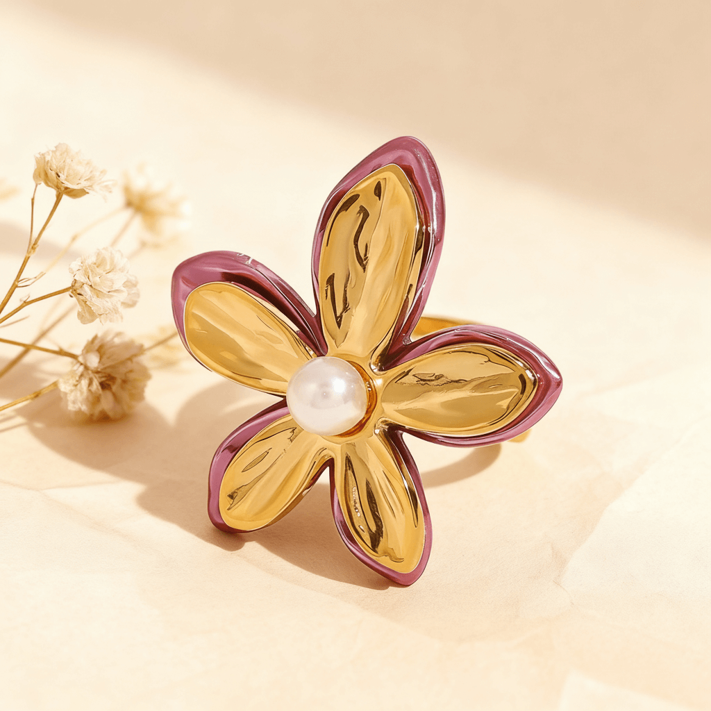 The Orchid Ring, a sculptural gold flower with fuchsia-pink enamel edges and a central white pearl, displayed on a white surface with a rose quartz crystal in the background. The ring is an open, adjustable cuff design.