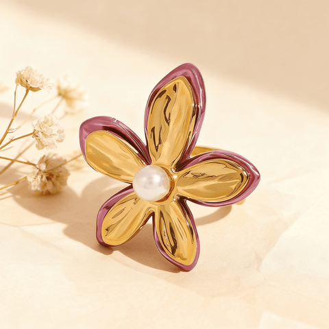 The Orchid Ring, a sculptural gold flower with fuchsia-pink enamel edges and a central white pearl, displayed on a white surface with a rose quartz crystal in the background. The ring is an open, adjustable cuff design.