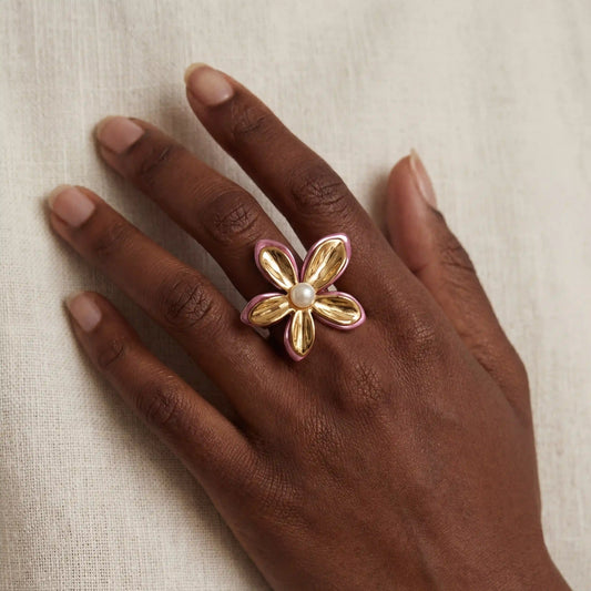 Close up of woman with dark skin wearing large gold orchid ring with pink enamel and pearl.