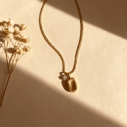 The Signature Necklace, featuring a rectangular brushed gold tag pendant and a small, asymmetrical CZ crystal charm on a delicate gold chain, displayed on a clean white background.