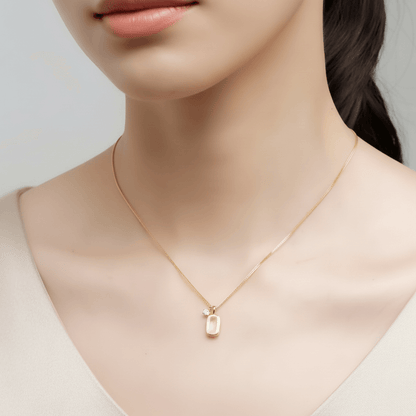 A close-up of a model's neck and collarbone, showcasing The Signature Necklace. The brushed gold tag rests elegantly in the center, while the small, sparkling CZ accent sits slightly off to the side, highlighting the piece's unique asymmetrical design.
