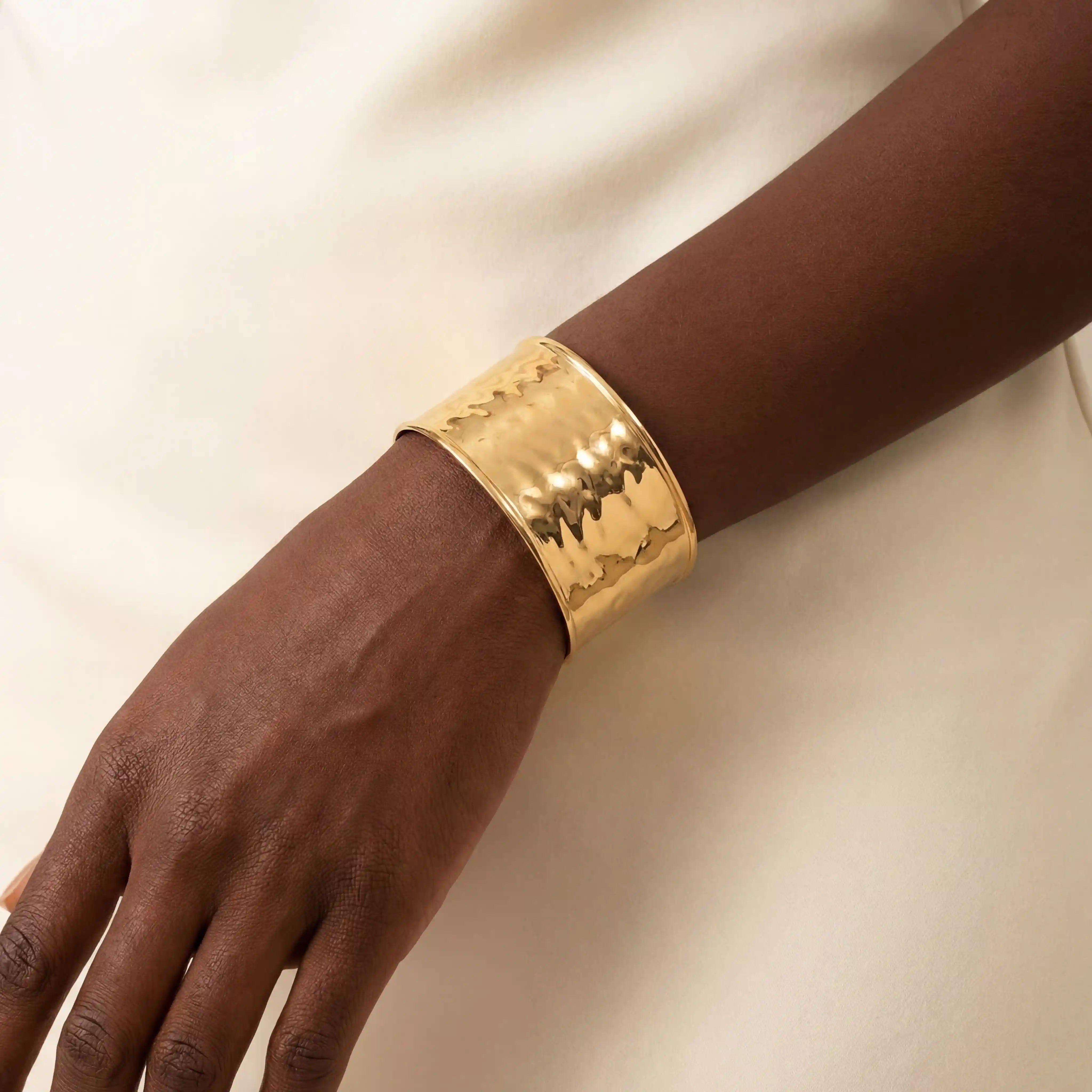 Close up of woman with dark skin wearing wide hammered gold cuff bracelet statement jewelry. Main image