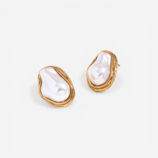 A detailed product shot of The Theia Earrings by Luminova, showcasing the unique, irregular baroque pearls cradled in sculptural, textured gold stud settings on a white silk background.