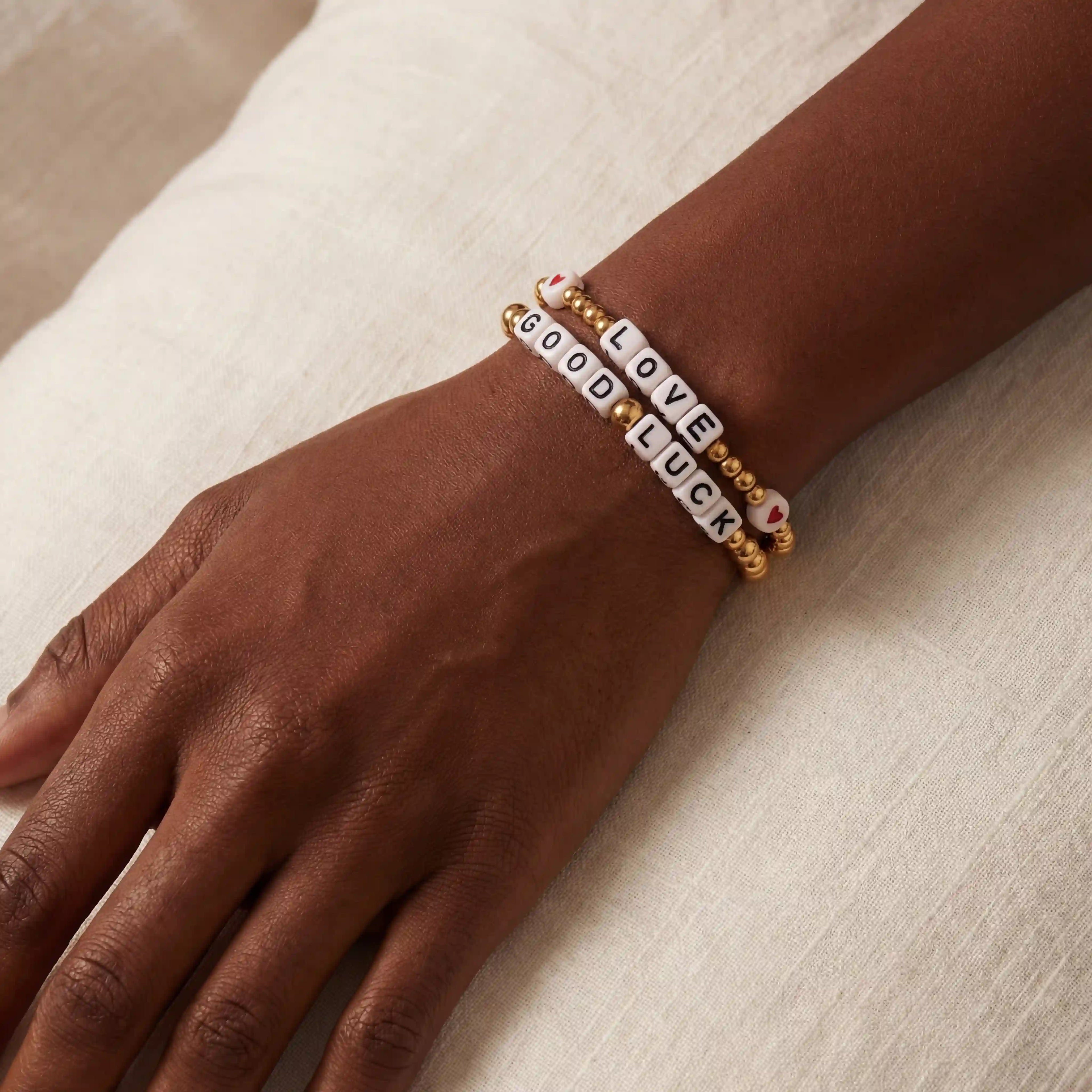 Close up of dark skin model wearing stacked gold beaded letter bracelets spelling LOVE and GOOD LUCK.