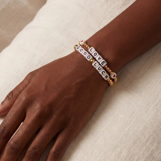 Close up of dark skin model wearing stacked gold beaded letter bracelets spelling LOVE and GOOD LUCK.