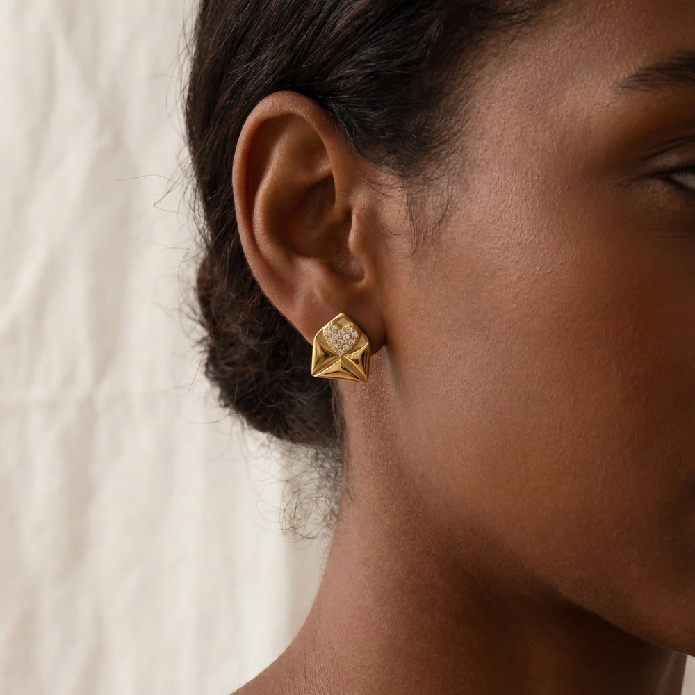 Close up of woman with dark skin wearing gold envelope stud earrings with crystal heart detail.