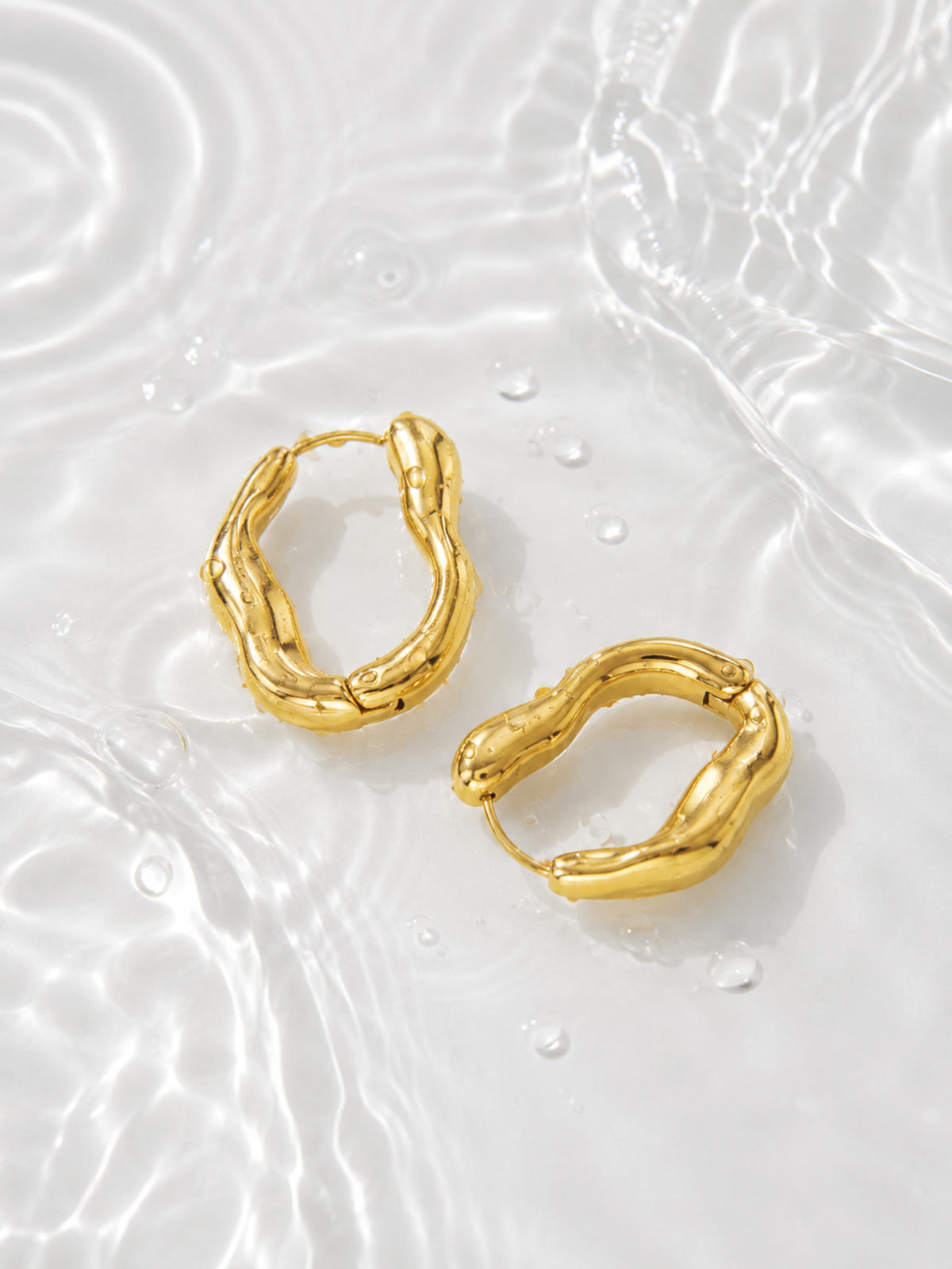 Sculpted Wave Huggie Hoop Earrings with water droplets on white water ripple background