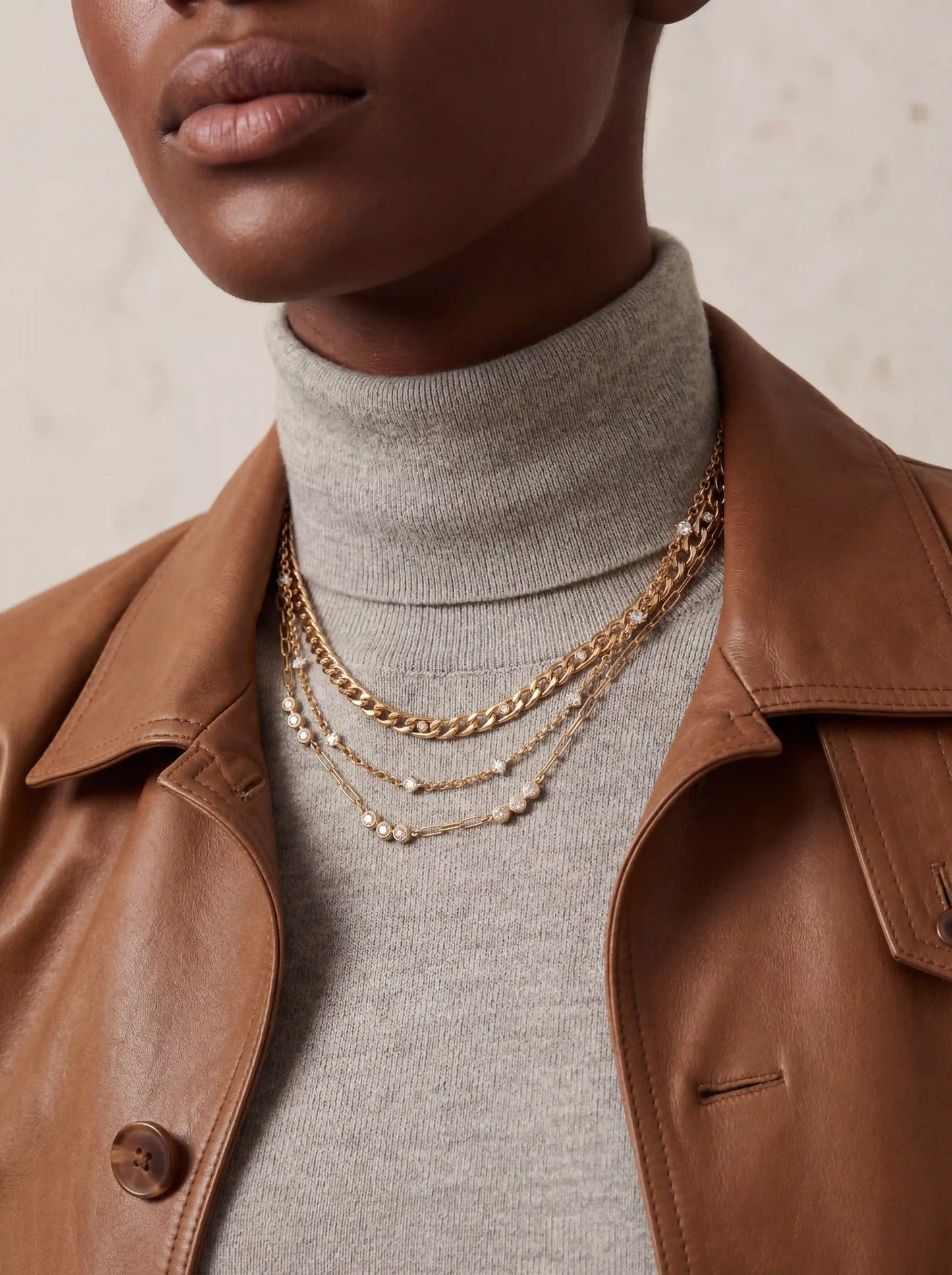 Woman styling bold gold layered necklace set over grey turtleneck and leather jacket fall fashion.