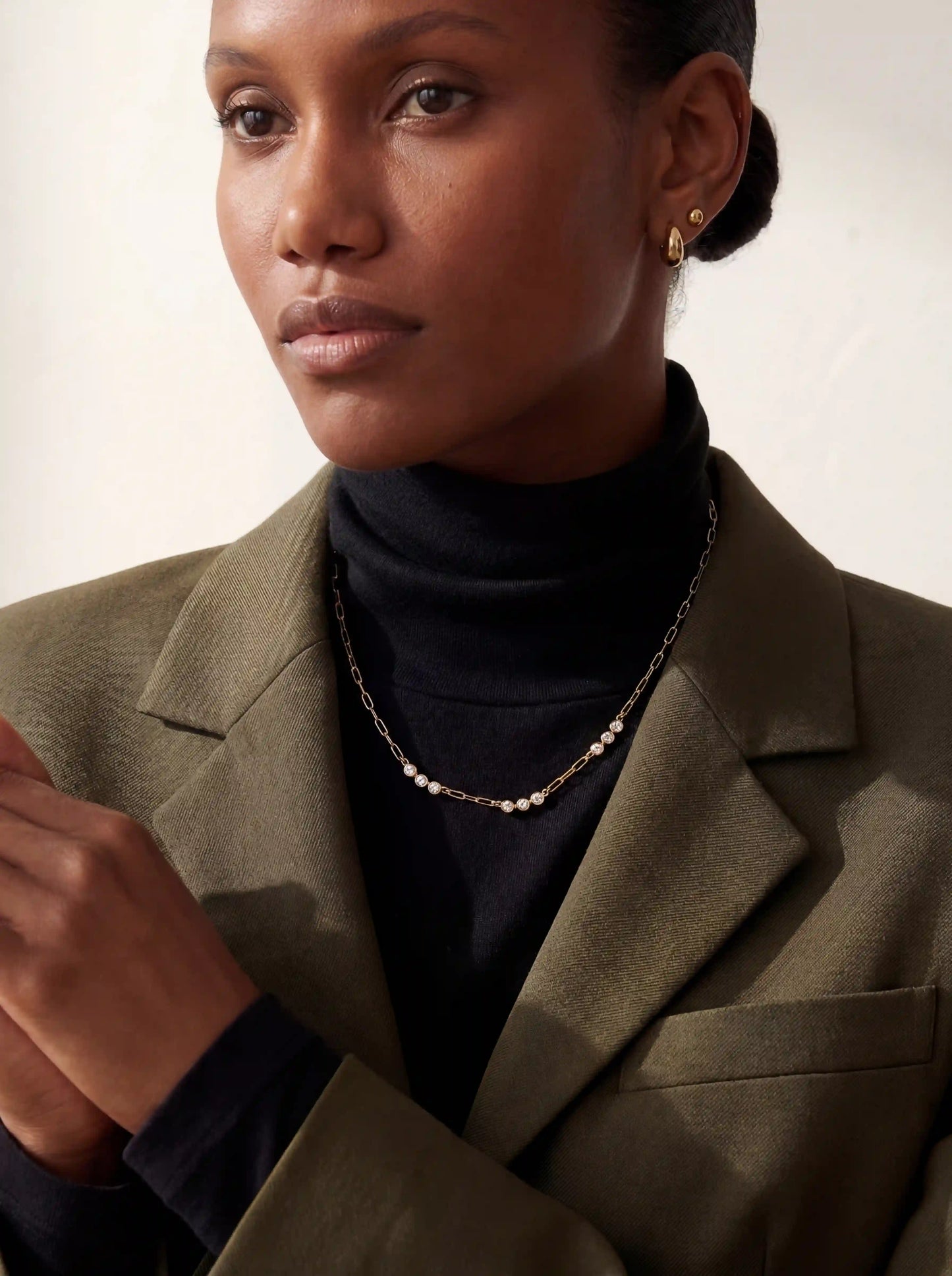 Model with dark skin wearing gold Trinity crystal necklace with olive blazer elegant office jewelry.