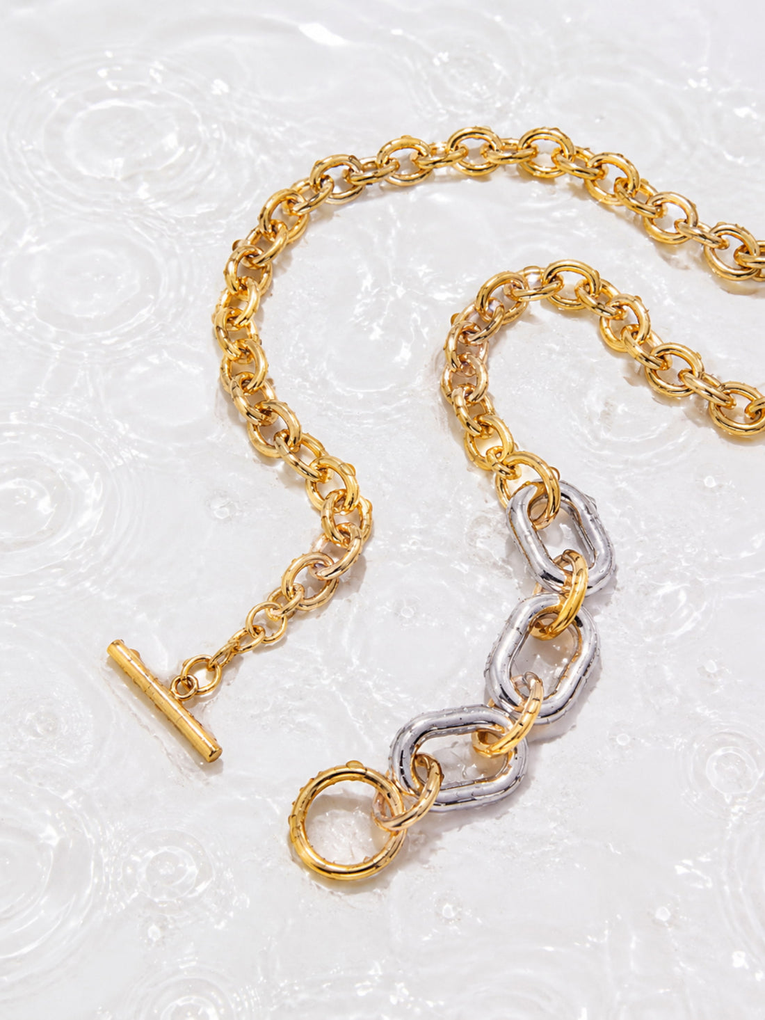 Two-tone chunky chain necklace with gold and silver links on a white water ripple background