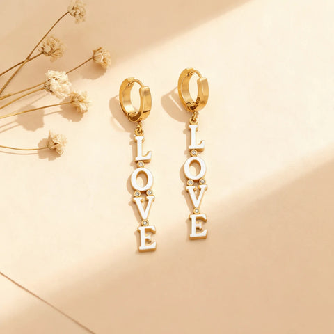 The Amor Earrings laid out on a soft, textured background, showcasing the graphic design of the 'LOVE' letter drops and the sparkle of the crystal accents.
