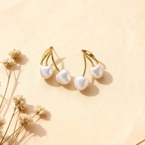 A pair of The Chérie Pearl Earrings by Luminova, styled on a soft beige background with delicate dried flowers, showcasing the cute cherry design with heart-shaped pearls.