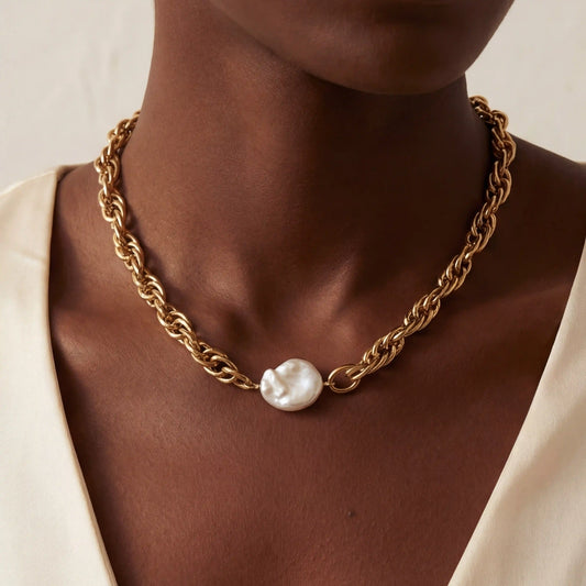 Close up of woman with dark skin wearing chunky twisted gold chain necklace with large baroque pearl center.