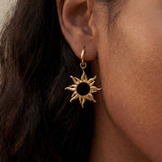 Close up of woman with dark skin wearing gold radiant sun charm drop earrings celestial jewelry.
