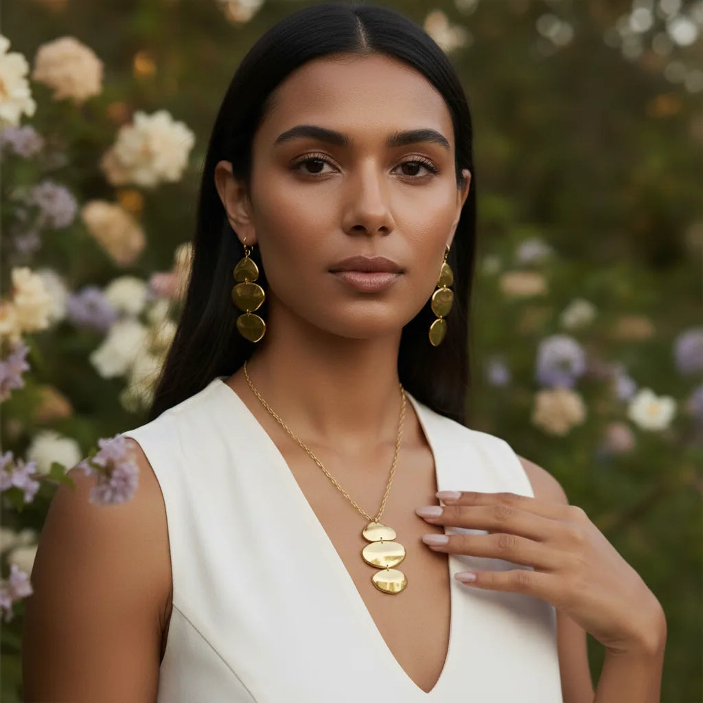 A model in a garden setting wearing The Thalia Earrings and a matching sculptural gold pendant necklace, showcasing a complete, artistic jewelry set.