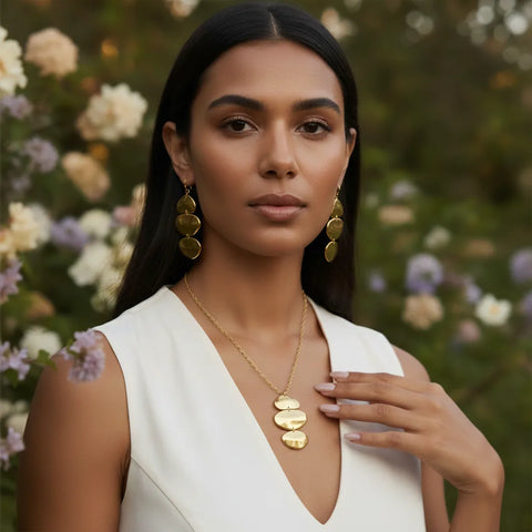 A model in a garden setting wearing The Thalia Earrings and a matching sculptural gold pendant necklace, showcasing a complete, artistic jewelry set.