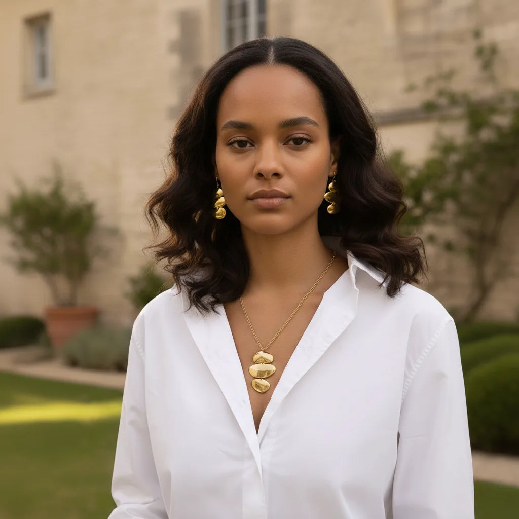 A beautiful model in a garden setting, touching her neck to feature both the sculptural Thalia Necklace and the matching Thalia Earrings.