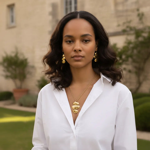 A beautiful model in a garden setting, touching her neck to feature both the sculptural Thalia Necklace and the matching Thalia Earrings.