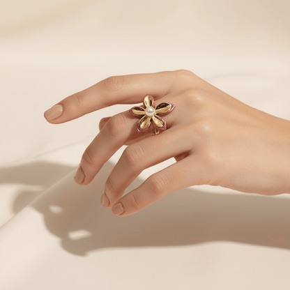 A close-up of The Orchid Ring on a model's hand in soft, natural light. The light catches the high-polish gold petals, the glossy pink enamel, and the soft luster of the central pearl, highlighting its exquisite details.