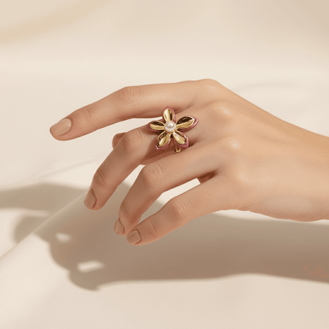 A close-up of The Orchid Ring on a model's hand in soft, natural light. The light catches the high-polish gold petals, the glossy pink enamel, and the soft luster of the central pearl, highlighting its exquisite details.