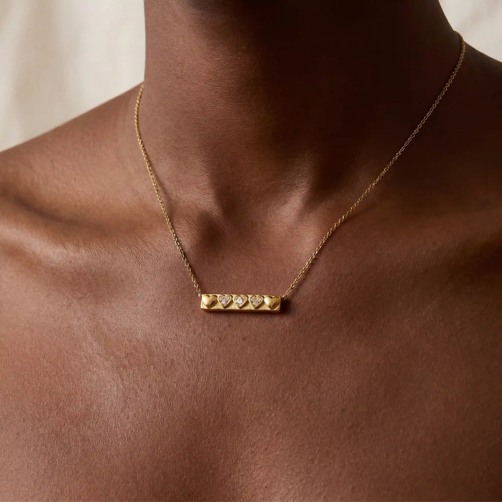 Close up of woman with dark skin wearing gold bar necklace with row of heart details.